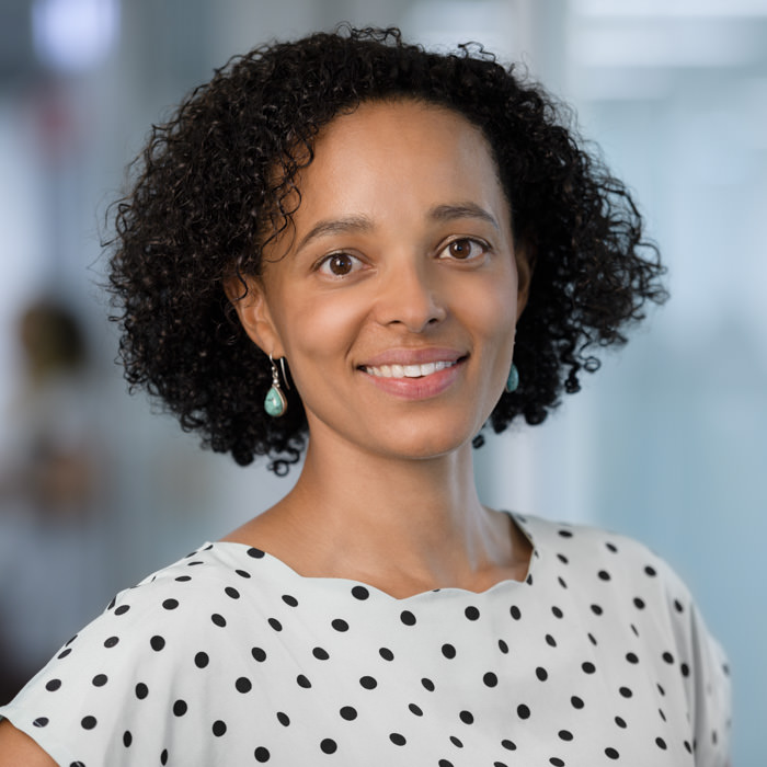 Professional headshot photography of a businesswoman in a Sydney office A professional headshot photograph of a smiling businesswoman with curly hair, wearing a polka dot blouse and turquoise earrings, captured in a modern Sydney office setting.