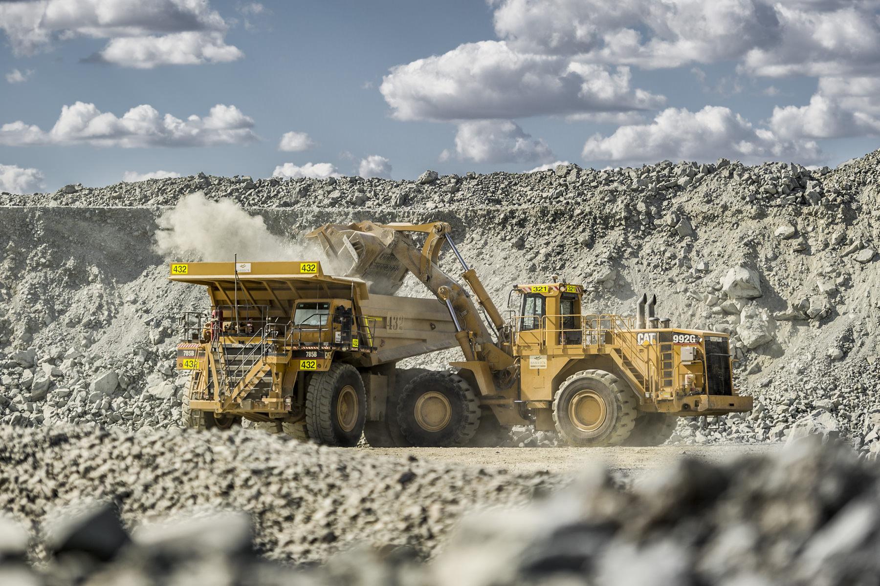Industrial mining photography of heavy machinery at an Australian quarry An industrial photograph of heavy machinery in action at an Australian quarry, showcasing a large front-end loader dumping rocks into a mining dump truck under a bright sky with scattered clouds.