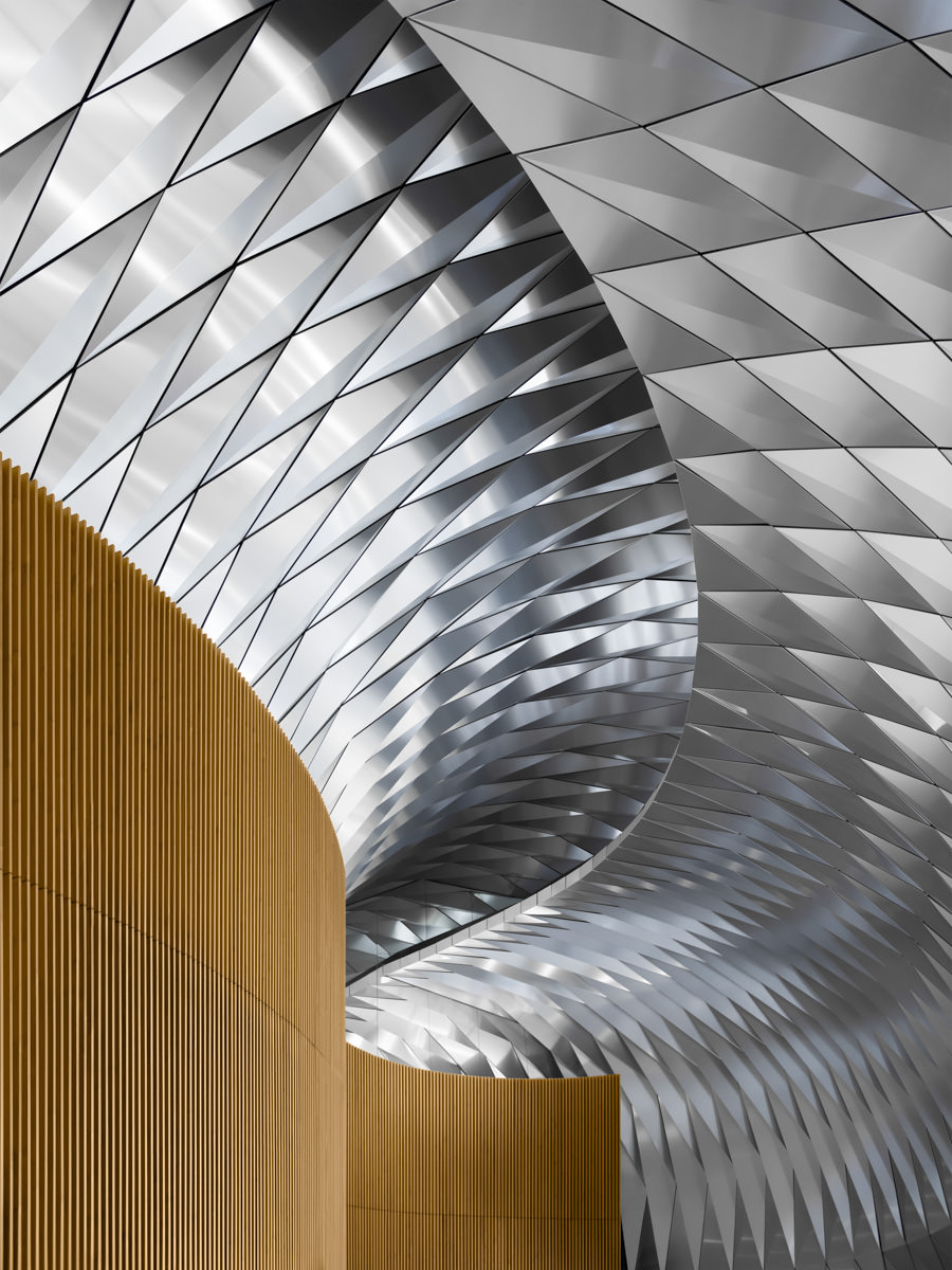 Interior architectural photography of a modern ceiling design in Sydney A detailed interior architectural photograph showcasing a dynamic metallic ceiling with intricate geometric patterns, contrasted by curved wooden paneling in a modern Sydney building.